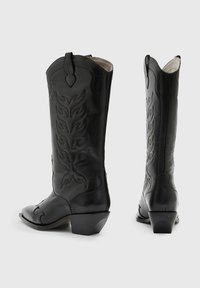 Svarta lädercowboystövlar med spetsig tå, dekorativt sydda mönster och en kort klossad klack. Har en dragrem för enkel påtagning.