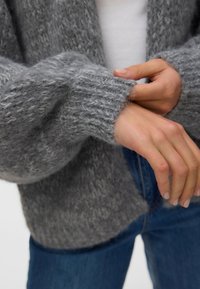 Άτομο που διορθώνει τη μανσέτα ενός γκρίζου, μαλακού πλεκτού πουλόβερ, φορώντας μπλε τζιν και άσπρο πουκάμισο από κάτω.