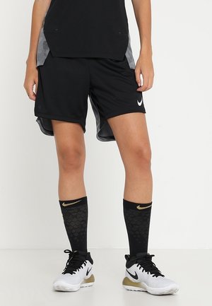 Personne portant un short de sport noir, des chaussettes noires hautes avec un logo Nike doré, et des chaussures de sport Nike blanches avec des lacets noirs.