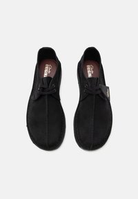Black suede shoes with a lace-up design, featuring smooth texture, rounded toe, and stitching accents. Includes a brand tag on one shoe.