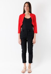 Giacca rossa corta con maniche a tre quarti sopra una canottiera nera, abbinata a pantaloni neri a sigaretta e sandali nudi con cinturini.
