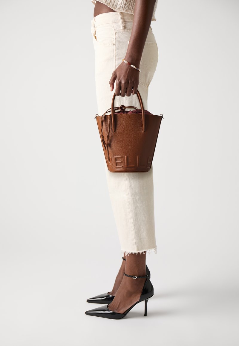 Brown leather bucket bag with embossed logo, short handles, and a drawstring closure, paired with white cropped jeans and black heels.