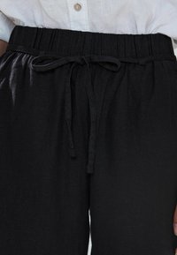 A fekete len nadrág rugalmas derékkal és húzózsinórral rendelkezik, sima textúrájú és laza szabású, világos gombos inggel párosítva.