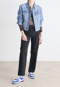 Persona vistiendo chaqueta de mezclilla azul claro sobre blusa negra transparente, jeans negros de tiro alto y zapatillas blancas con detalles en azul, de pie sobre suelo beige.