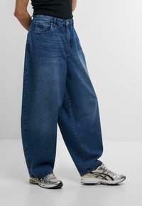 Pantaloni in denim scuro e larghi con cinque tasche, cuciture visibili e chiusura a bottone tradizionale. Abbinati a sneakers grigie e argentate.