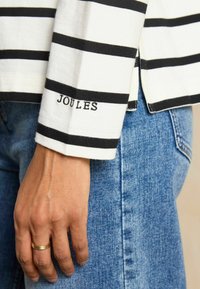 Langärmliges Shirt mit weißen und schwarzen Streifen, mit "JOULES" auf dem Manschetten bestickt. Getragen mit blauen Jeans, was einen lässigen Stil zeigt.