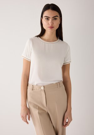 Femme aux cheveux lisses et foncés portant une chemise blanche à manches courtes avec une bordure beige et un pantalon beige taille haute, debout devant un fond clair.