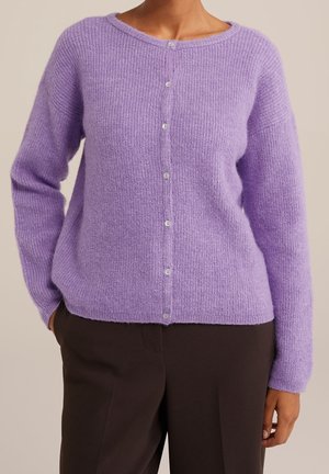 Persona que lleva un cárdigan de punto lavanda abotonado con mangas largas y pantalones marrón oscuro sobre un fondo liso.