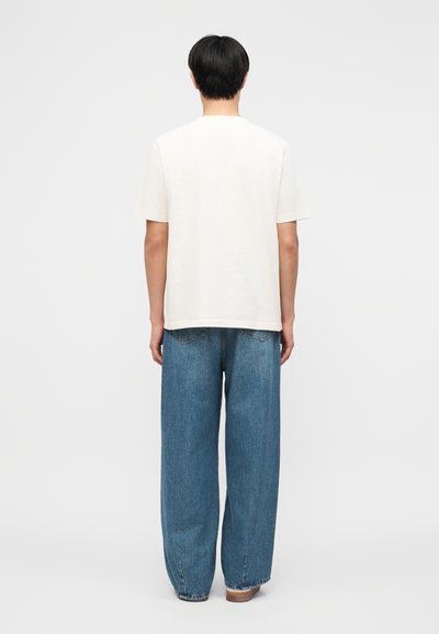 Άτομο που στέκεται με την πλάτη, φορώντας απλό λευκό κοντομάνικο μπλουζάκι και φαρδύ μπλε τζιν, μπροστά σε απλό φόντο.