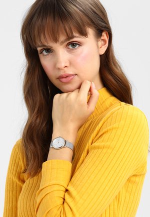 Joven mujer con cabello castaño que lleva un suéter amarillo con textura de canalé y un reloj de pulsera plateado, apoyando su barbilla sobre su mano con fondo blanco.