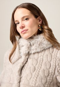 Cappotto in pelliccia sintetica grigia con motivo a maglia strutturata, caratterizzato da un colletto morbido e soffice. Orecchini dorati con accenti colorati completano il look.