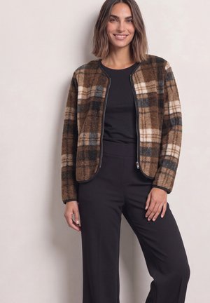 Femme portant une veste zippée à carreaux marron et beige sur un haut noir et un pantalon noir, debout et souriante devant un fond uni.