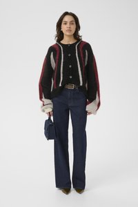 Frau in einem schwarz-rot-grauen gestreiften Knopfpullover, dunklen blauen Jeans, olivfarbenen spitzen Schuhen und mit einer kleinen navy Handtasche.
