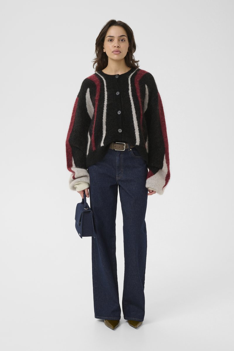 Donna che indossa un maglione a righe nero, rosso e grigio con bottone, jeans blu scuro, scarpe a punta oliva e porta una piccola borsa a tracolla blu navy.