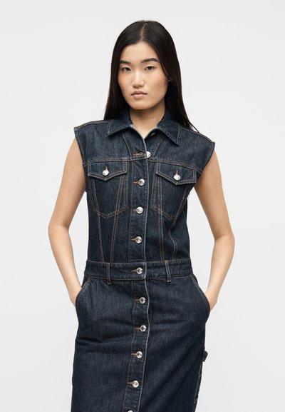 Γυναίκα με μακριά σκούρα μαλλιά, φορά ένα αμάνικο σκούρο τζιν φόρεμα με κουμπιά και μπροστινές τσέπες, με τα χέρια στις πλαϊνές τσέπες.