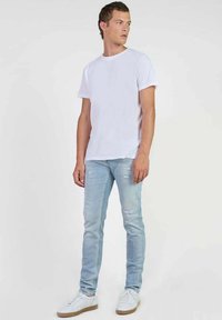 Camiseta blanca de algodón combinada con jeans skinny de algodón ligeros y desgastados en azul claro, y zapatillas blancas con suela beige, de pie contra un fondo liso.