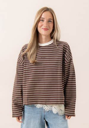 Junge Frau mit langen blonden Haaren, die ein braun-weiß gestreiftes langärmeliges Shirt über einem Spitzenbesetzen Top und hellblauen Jeans trägt und lächelt.