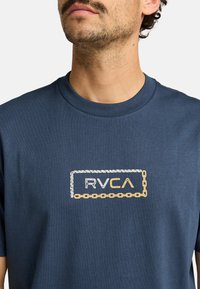 Camiseta de algodón azul marino con un gráfico rectangular que presenta "RVCA" y un diseño de cadena en dorado y blanco. Estilo clásico de cuello redondo.