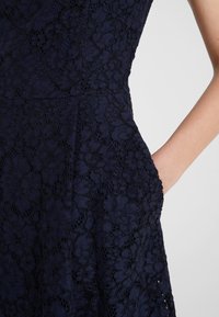 Robe en dentelle bleu marine à motif floral, taille ajustée et main reposant dans la poche latérale sur fond uni.