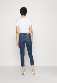 Jeans ajustados azules de cintura alta, con un diseño clásico de cinco bolsillos y un sutil detalle de costura, combinados con una camiseta blanca y tacones blancos.