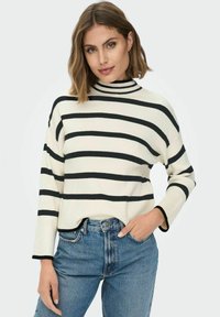 Svītrains džemperis krēmkrāsā un melnā krāsā ar augstu apkakli, ribota tekstūra, nolaistas plecu līnijas un brīva piegriezuma. Kombinēts ar zilām džinsu biksēm.