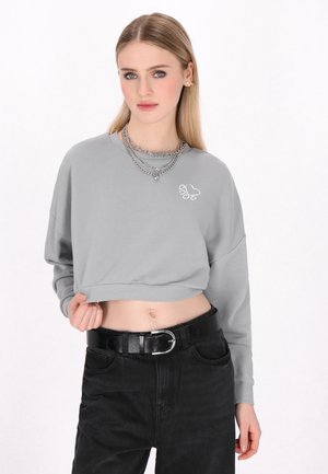 Harmaa lyhyt hiha sweatshirt, jossa on pitkät hihat ja valkoinen tassunjälki-logo. Yhdistetty korkeavyötäröisten mustien farkkujen ja mustan vyön kanssa.