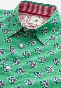 Grüne Druckknopfbluse mit floralem Muster, das pinke und weiße Blumen zeigt. Innenkragen mit rot-weißen Streifen. Aus Baumwollmaterial.