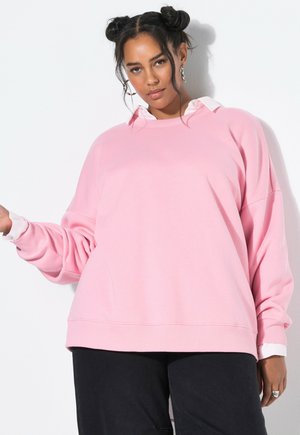 Donna con due chignon che indossa una felpa rosa oversize sopra una camicia bianca e pantaloni neri, in piedi contro un muro bianco.