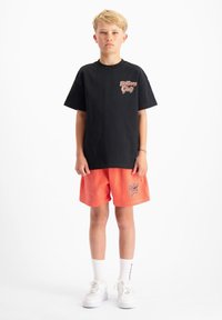 Zwarte katoenen T-shirt met korte mouwen en een logo op de voorkant, gecombineerd met koraalkleurige shorts met hetzelfde logo, en witte sneakers.