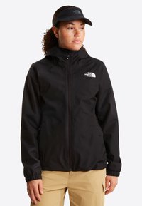 Γυναίκα με μαύρο μπουφάν North Face και μαύρη καπέλο-visor, που κοιτάει προς το πλάι, όρθια μπροστά σε απλό λευκό φόντο.
