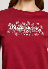 Röd bomullströja med blommönster som har "Pepe Jeans" i kursiv och "LONDON" i fet stil nedanför. Kort ärm, rund halsringning.