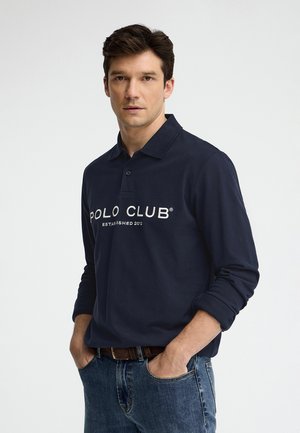 Hombre con cabello oscuro que lleva una camisa polo azul marino de manga larga con el texto “POLO CLUB Established 2012”, manos en los bolsillos del vaquero, fondo neutro.