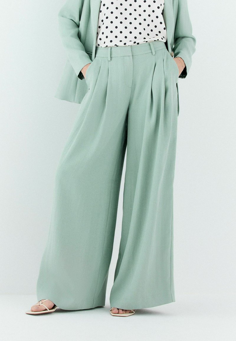 Femme portant un pantalon large vert sauge, un blazer assorti, un chemisier à pois noir et blanc et des sandales blanches à bout carré, les mains dans les poches.