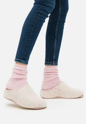 Beige Filzpantoletten mit glatter Textur, runder Form und hellrosa gerippten Socken. Getragen mit dunkelblauen engen Jeans, die den Sockenbund zeigen.