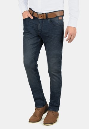Jeans in denim blu scuro con vestibilità slim, dotati di tasca frontale e una cintura in pelle marrone con fibbia metallica e dettagli con etichetta.