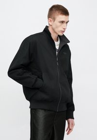 Μαύρο bomber μπουφάν με ψηλό γιακά, κλείσιμο με φερμουάρ, πλαϊνές τσέπες και ριμπ μανσέτες. Μαλακό ύφασμα με διακριτικά στοιχεία στα μανίκια.