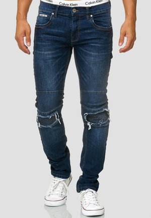 Vaqueros de mezclilla azul oscuro con un corte ajustado, detalles de desgaste en las rodillas y una cinturilla de Calvin Klein. Combinados con zapatillas deportivas blancas.
