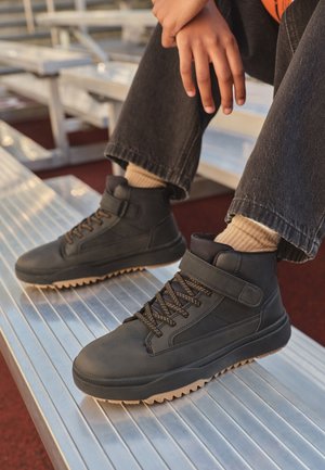 Personne portant des baskets montantes noires avec des semelles beiges, des chaussettes beiges et un pantalon sombre, assise sur des gradins métalliques en plein air.