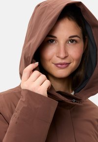 Veste imperméable marron avec capuche, dotée d'une fermeture éclair à l'avant et d'une texture lisse. La manche gauche présente un détail de logo imprimé.