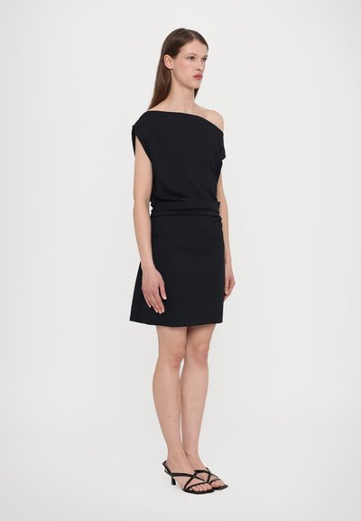 Zwarte off-the-shoulder jurk met een aansluitend lijfje en een uitlopende rok. Gemaakt van een gladde stof. Gecombineerd met zwarte sandalen met hakken en banden.