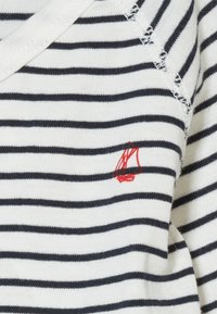 Tessuto bianco con strisce orizzontali navy, caratterizzato da un piccolo logo di una barchetta a vela ricamata in rosso. Design a V con cuciture testurizzate.
