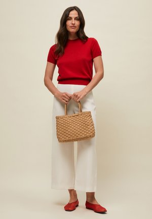 Žena v červenom tričku s krátkym rukávom a bielych širokých nohaviciach drží pletenú béžovú kabelku, stojí pred béžovým pozadím.