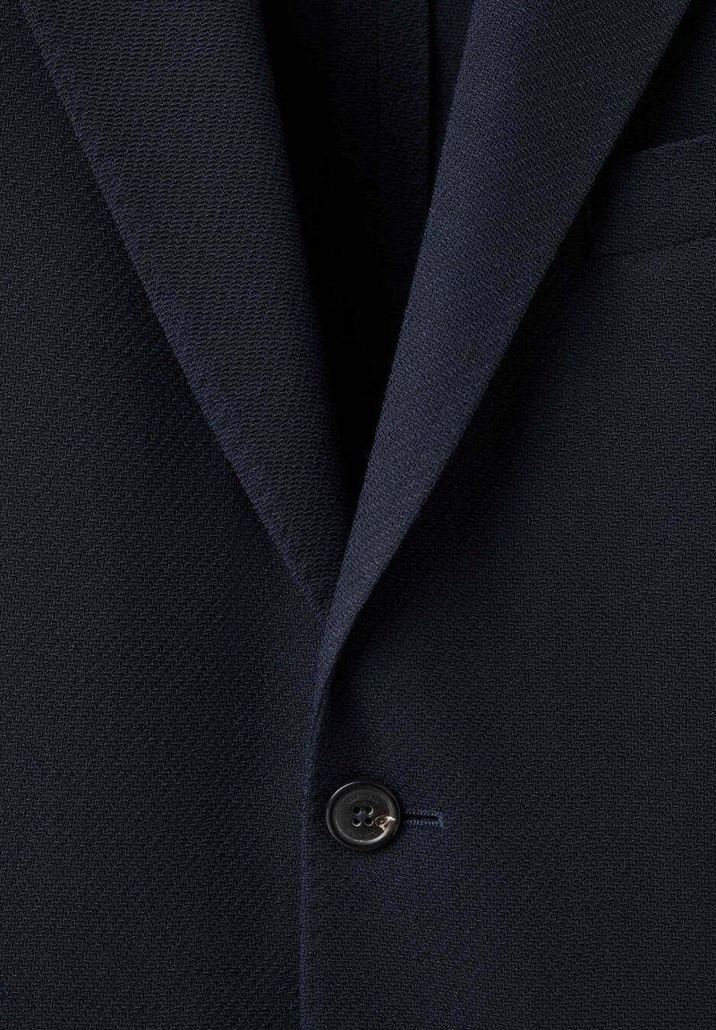 Blazer azul marino oscuro con tela texturizada, solapa con muesca, un solo botón negro abrochado y un bolsillo en el pecho en el lado derecho.
