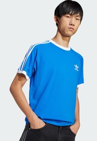 Blue cotton t-shirt with white ribbed collar and sleeve cuffs, featuring three white stripes on shoulders and a small white logo on chest.
