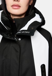 Schwarze und weiße Jacke mit hohem Kragen, Reißverschluss vorne und Kontrastpanelen. Ausgestattet mit gebrandeten Textilien und metallischen Akzenten.