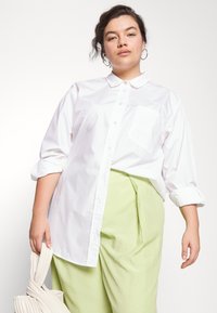 Camisa branca de colarinho com botões e mangas longas, com um bolso no peito. Combinada com umas calças plissadas de verde claro e uma bolsa bege.
