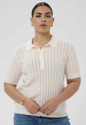 Kobieta o ciemnych włosach, ubrana w biały, krótki sweterkowy polo, niebieskie dżinsy, złote kolczyki koła i złotą bransoletkę, pozująca z rękami złączonymi.