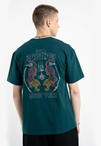 Mężczyzna w ciemnozielonej koszulce z kolorową grafiką tygrysa i napisem "Muay Thai", widoczny od tyłu na białym tle.