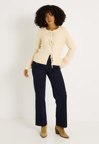 Cárdigan beige de punto con cierre frontal de lazo, con detalles rosas en las mangas. Combinado con jeans de pierna ancha azul oscuro y botines marrón claro.