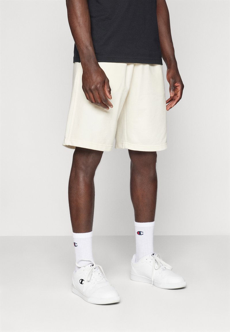 Weiße Fleece-Shorts mit lockerer Passform, kombiniert mit weißen Sneakers und Socken mit Logo. Das Oberteil ist ein einfarbiges schwarzes T-Shirt. Keine Muster.